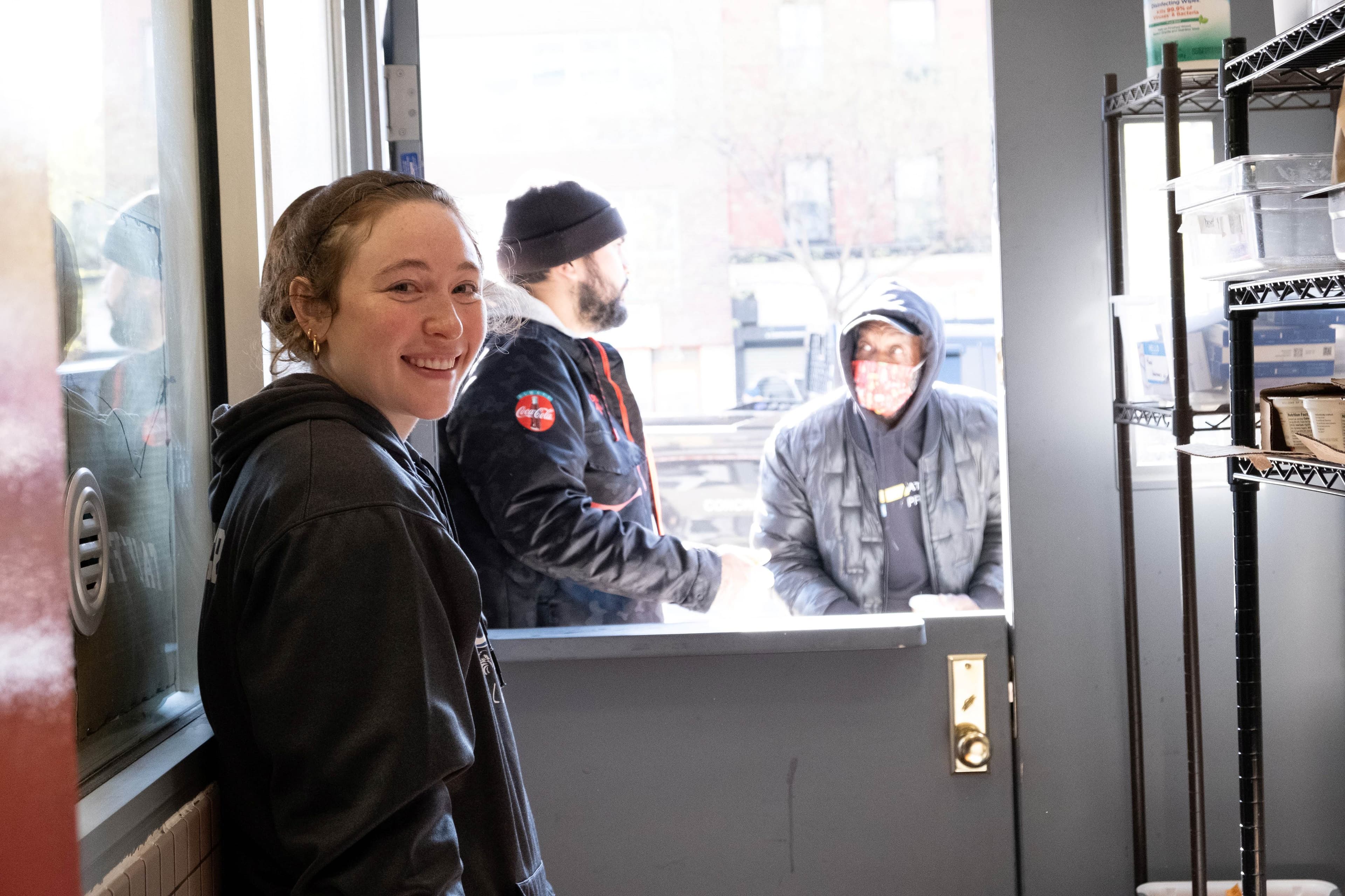 One woman staring at the camera smiling. Two people are outside a service window looking at one another.